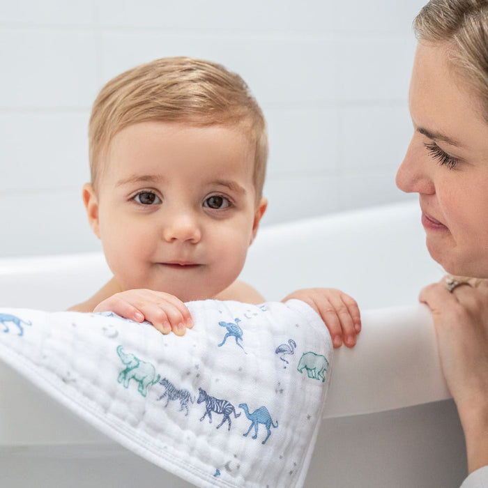 A baby with short blonde hair sits in a bathtub, holding an aden + anais boutique 100% cotton muslin oversized washcloth featuring animal prints and absorbent layers. A light-haired woman smiles warmly at the baby against a plain, light-colored wall background.