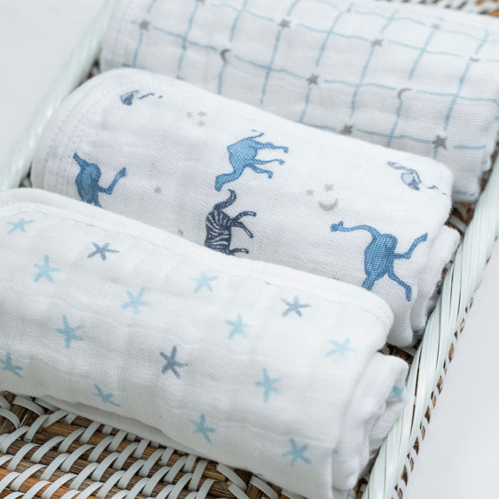 Three boutique 100% cotton muslin oversized washcloths by aden + anais sit in a wicker basket. Each washcloth features unique designs: blue stars, cartoon animals, and a blue grid with animal motifs. Their absorbent layers ensure comfort and warmth for your baby’s sensitive skin.