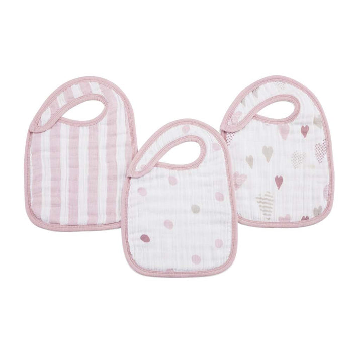 The aden + anais boutique 100% cotton muslin snap bibs 3 pack includes three absorbent bibs with soft pink trim: one with stripes, another with dots, and the third adorned with hearts and arrows on a white background, all featuring an adjustable fit for comfort.