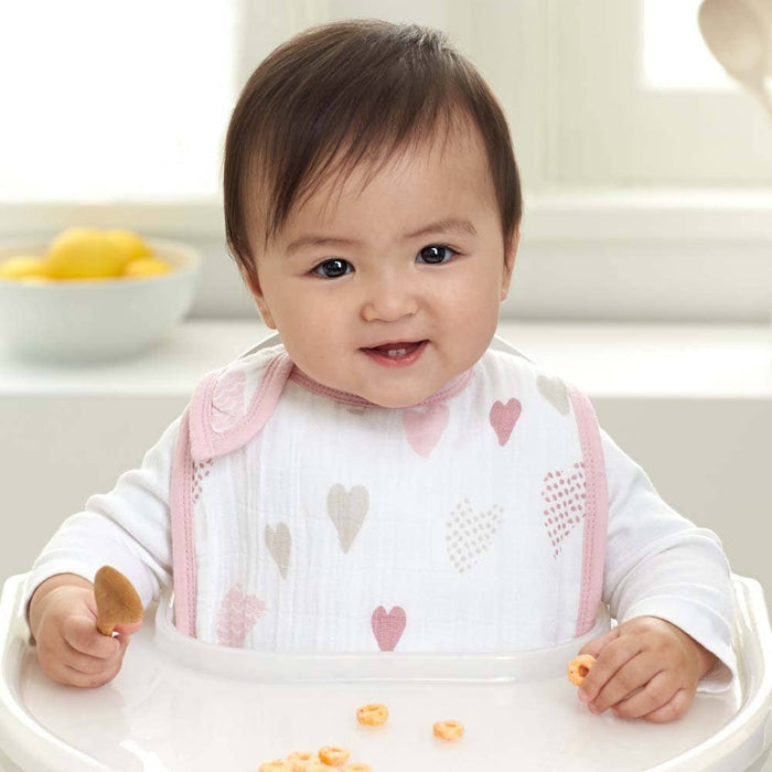 A smiling baby sits in a high chair wearing an aden + anais boutique 100% cotton muslin snap bib adorned with pink and gray hearts, comfortably holding a cereal-topped spoon. A bowl of lemons brightens the background on a table by the window.