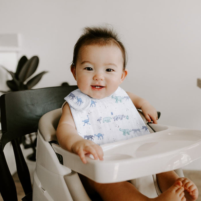 A smiling baby sits in a high chair, wearing Snap Bibs from the aden + anais Messy Babe Bundle with blue and green animal prints. A plant is partially visible to the left against a simple background, adding warmth.