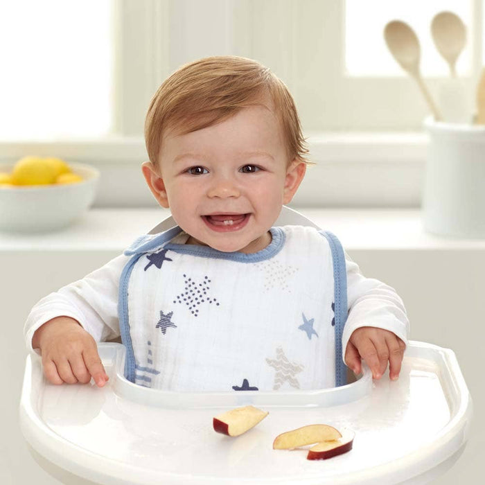 A baby beams in a high chair wearing an aden + anais boutique 100% cotton muslin snap bib with a star pattern. Sliced fruit is on the tray, and the background reveals a bright kitchen with a bowl of lemons and wooden utensils.