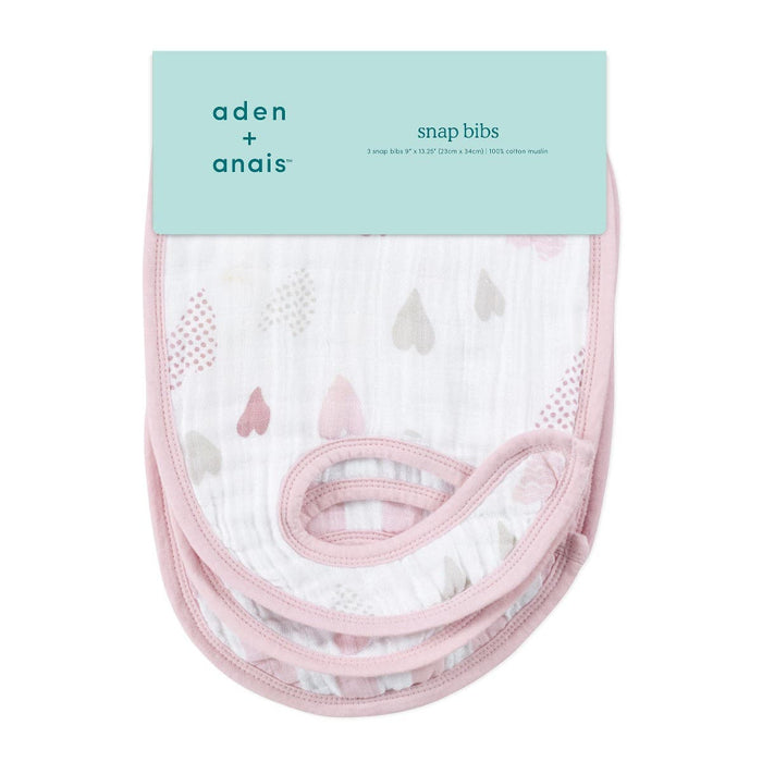 Three aden + anais boutique 100% cotton muslin snap bibs are stacked, showing their adjustable fit with packaging on top. The absorbent white bibs have light pink borders and a charming pattern of small pastel-colored hearts and dots.
