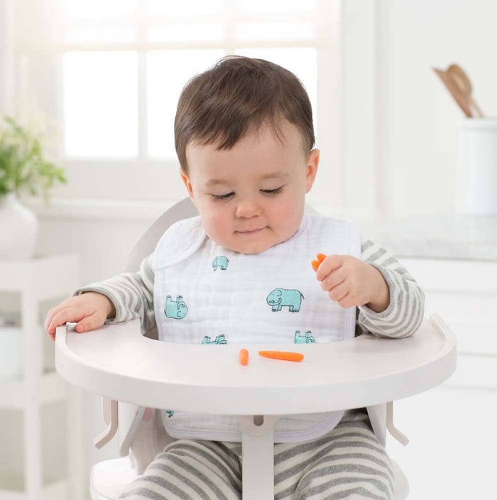 In a bright kitchen, a baby sits in a high chair wearing an aden + anais boutique 100% cotton muslin snap bib with animal prints for an adjustable fit. The baby is focused on a small carrot stick in their hand, with more carrot pieces on the tray.