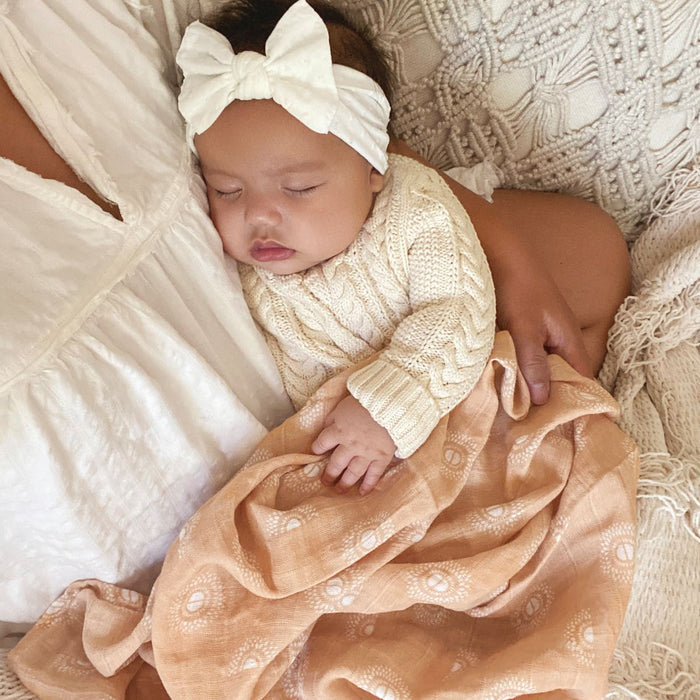 A baby in a cream sweater and white bow headband sleeps peacefully on a textured blanket and an adults arm, wrapped in an aden + anais boutique 100% cotton muslin swaddle blanket, featuring breathable peach fabric with white patterns.