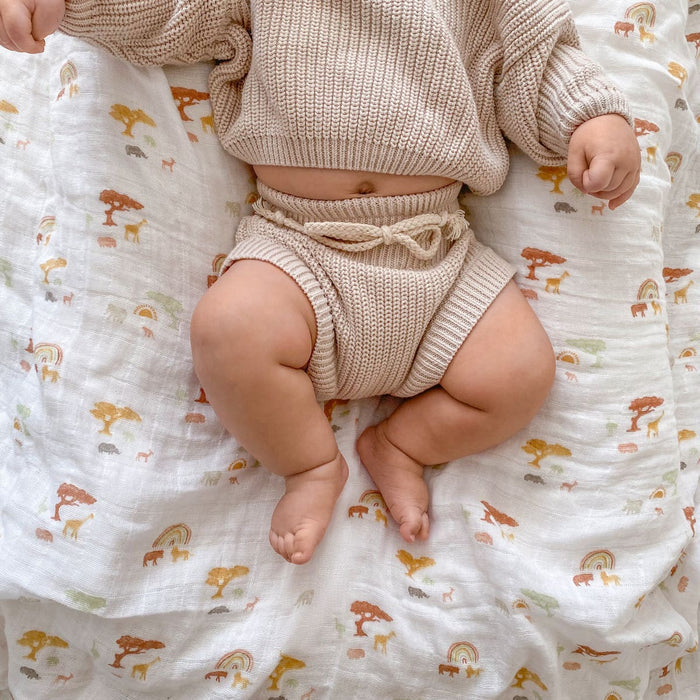 A baby rests on a soft, white blanket featuring animal and rainbow prints, wearing a cream knitted sweater and shorts. This scene showcases the comfort of aden + anais boutique 100% cotton muslin swaddle blankets, celebrated for their breathability and delightful textures.
