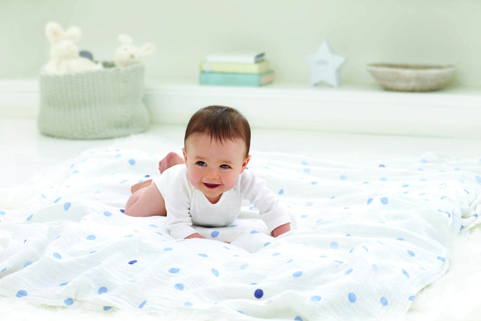 A baby in a white onesie lies on its stomach on an aden + anais boutique 100% cotton muslin swaddle blanket, featuring blue polka dots. The room’s soft, neutral palette includes a basket, books, and decor in the background. The baby is smiling.