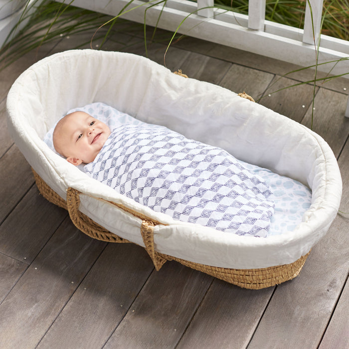 A baby smiles in a beige wicker bassinet, wrapped in a breathable aden + anais boutique 100% cotton muslin swaddle, set on a wooden floor near white railings and green plants.