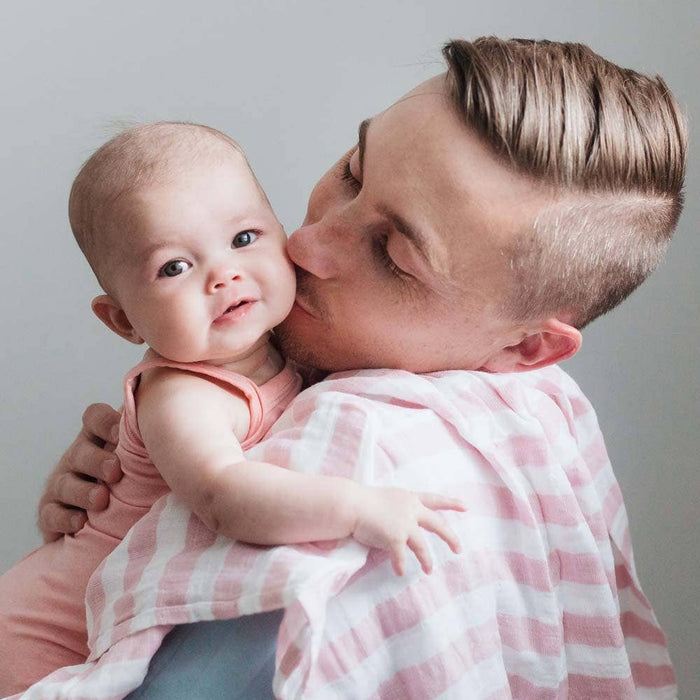 A man kisses a baby on the cheek while holding an aden + anais boutique 100% cotton muslin swaddle blanket over his shoulder. The baby, wearing a pink outfit, looks curiously at the camera against a plain, light-colored wall.
