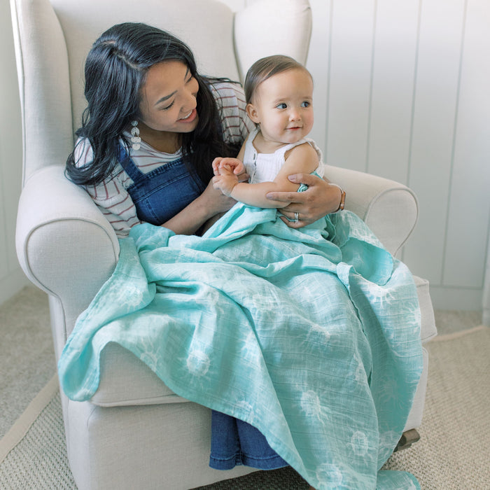A woman with long black hair cuddles her baby in a light armchair, using an aden + anais boutique 100% cotton muslin swaddle blanket with a turquoise and white design. Both smile warmly, enjoying their cozy room with light walls and carpet.