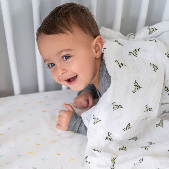A baby is lying on its stomach in a crib, smiling. The baby is partially covered with an aden + anais boutique 100% cotton muslin swaddle blanket from a 4 pack, and the crib sheet has multi-colored letters. The baby is wearing a gray outfit.