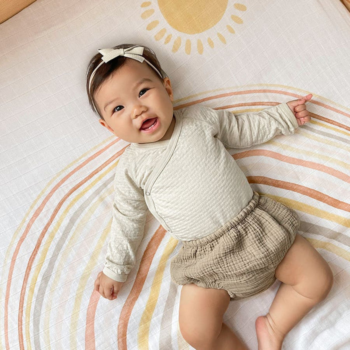 A smiling baby in a beige outfit and headband with a bow lies on an aden + anais boutique 100% cotton muslin swaddle blanket, known for its sun and rainbow designs.