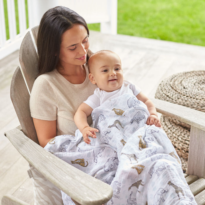 A woman sits on a wooden chair, holding a smiling baby wrapped in an aden + anais boutique 100% cotton muslin swaddle blanket with animal prints. They are on a porch with a woven circular rug and lush grass, enjoying their serene settings absorbency and comfort.