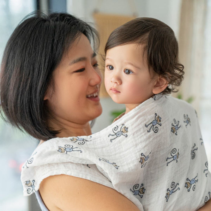An adult cradles a brown-haired child in a boutique 100% cotton muslin swaddle blanket by aden + anais, featuring cartoon monkeys. The adult smiles warmly while the child gazes away indoors, blurred windows softly framing their tender moment.