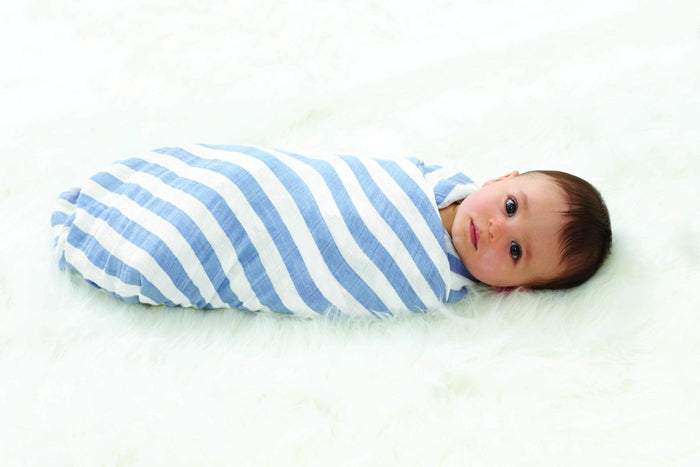 A baby lies comfortably in an aden + anais boutique 100% cotton muslin swaddle blanket, featuring blue and white stripes, on a soft white surface. The infants short brown hair and bright eyes enhance the cozy scene.