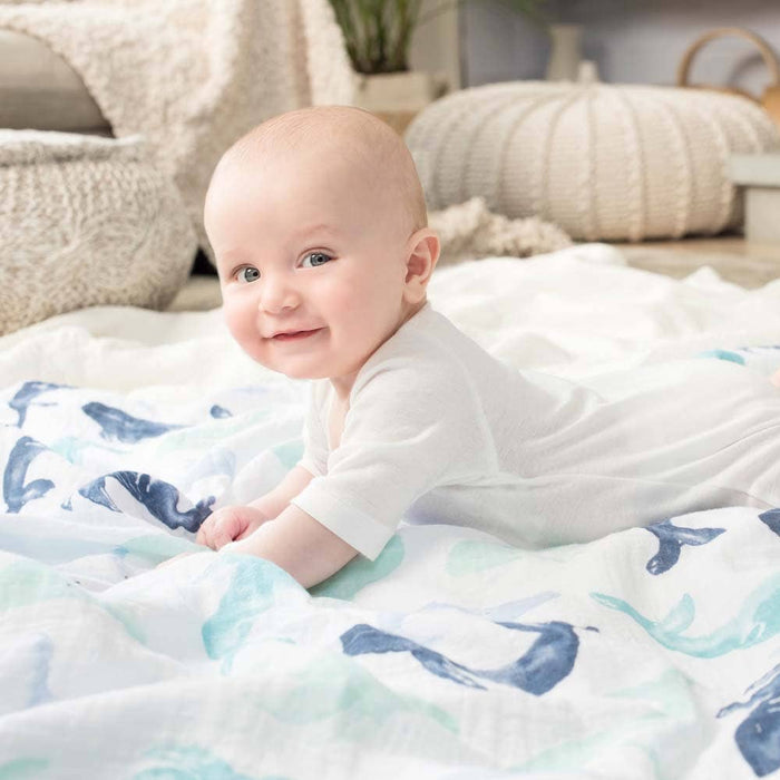 A smiling baby in a white outfit lies on an aden + anais boutique 100% cotton muslin swaddle blanket featuring a white and blue pattern. The background showcases soft furnishings that contribute to a cozy, serene atmosphere with muted colors.