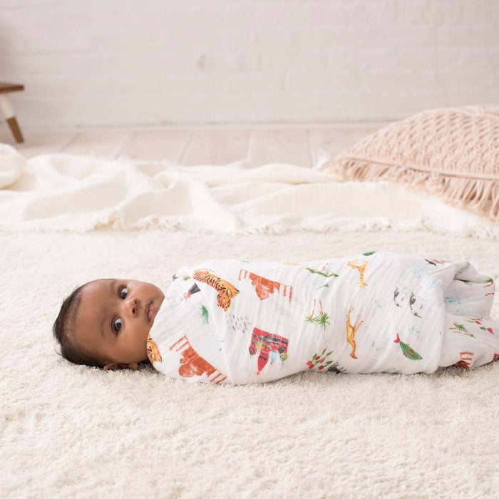 A baby rests enveloped in a breathable aden + anais boutique 100% cotton muslin swaddle blanket with colorful animal prints, lying on a plush light-colored blanket. The setting is accented with a textured pillow and extra blankets scattered over the warm wooden floor.