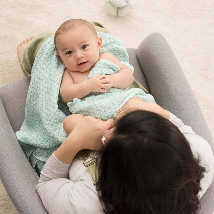 A baby wrapped in a light green aden + anais boutique 100% cotton muslin swaddle looks up while lying on the lap of an adult seated in a gray chair. The adults face is unseen, and a plush toy lies on the cream carpeted floor in the background.