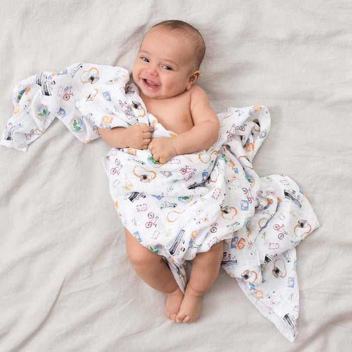 A smiling baby is wrapped in a breathable, colorful aden + anais boutique 100% cotton muslin swaddle blanket, lying on a cream-colored surface.
