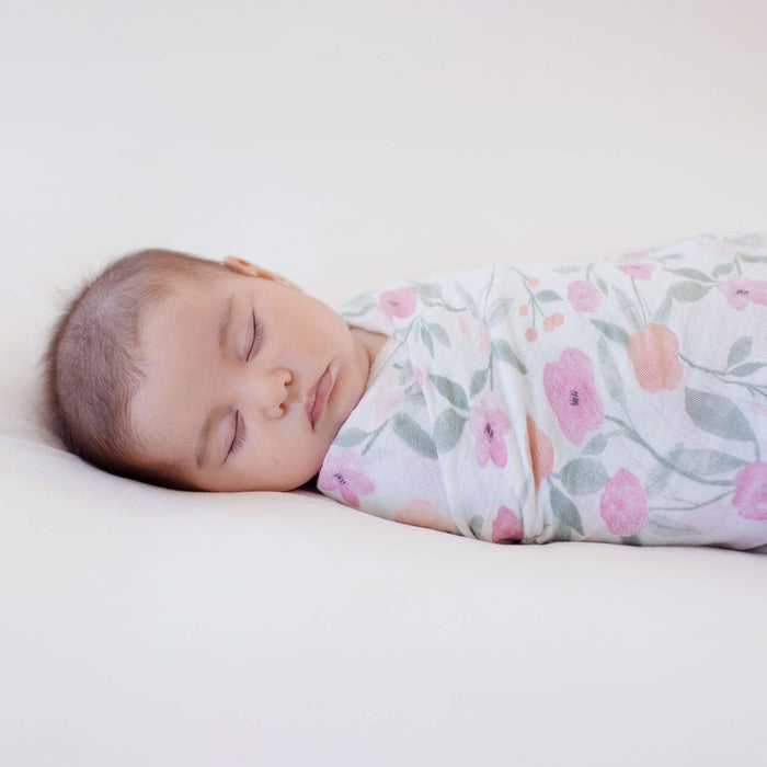 A sleeping baby wrapped in an aden + anais boutique 100% cotton muslin swaddle blanket with a floral pattern rests peacefully on a soft, white surface.