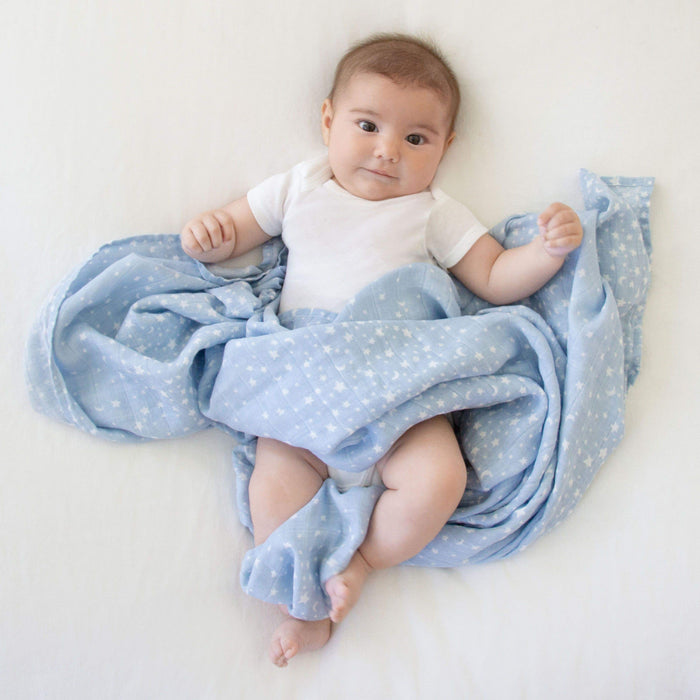 A baby lies on a white surface, wrapped in an aden + anais boutique 100% cotton muslin swaddle blanket with a light blue star pattern. The baby wears a white onesie and has a pleasant expression, with both arms slightly raised.