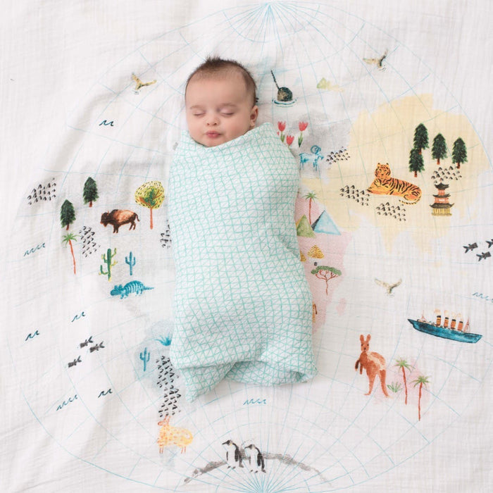 A baby peacefully sleeps, wrapped in an aden + anais boutique 100% cotton muslin swaddle. The breathable fabric gently surrounds the baby on a map-themed blanket, featuring animals, trees, and landmarks that create a whimsical scene.