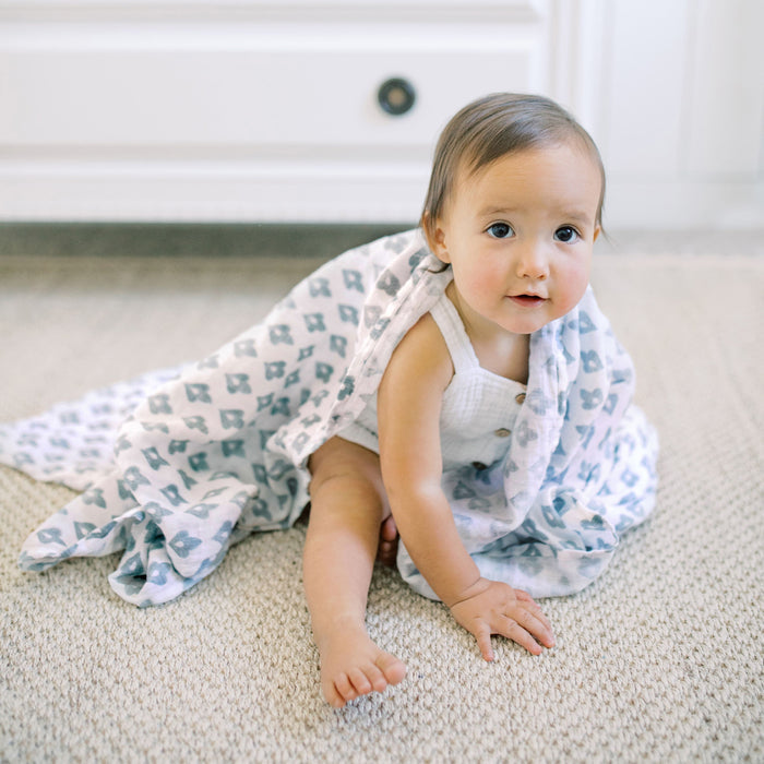 A baby sits on a carpeted floor, wrapped in a Messy Babe Bundle by aden + anais. Wearing a white sleeveless outfit, the baby curiously looks at the camera while a white drawer unit in the background completes the serene and cozy scene.