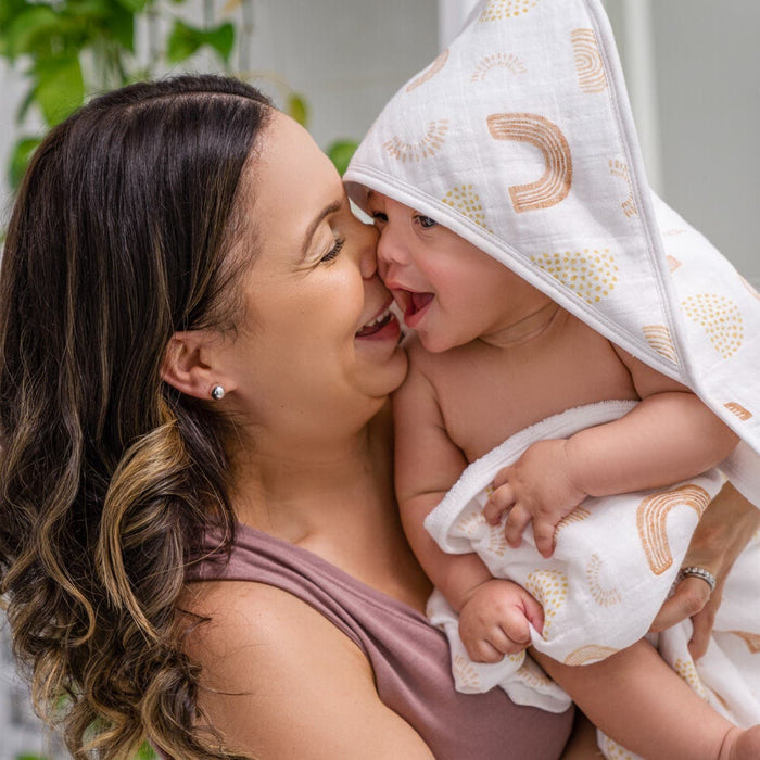 A woman happily holds a laughing baby wrapped in an aden + anais boutique 100% cotton oversized muslin-backed towel. They touch noses joyfully, with the woman in a sleeveless top and green plants visible behind them.