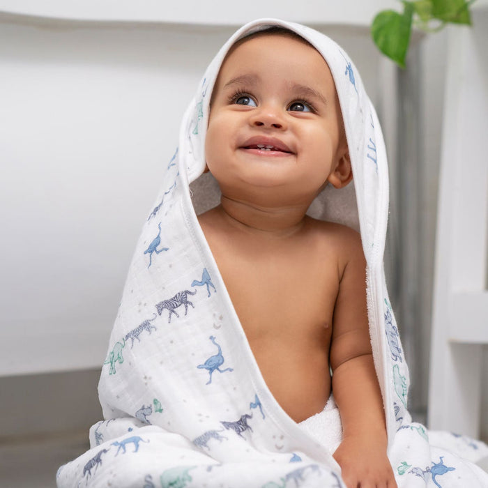 A happy baby sits upright, wrapped in the aden + anais boutique 100% cotton oversized muslin backed towel with animal prints, looking cozy and content. The hood gently covers the babys head, while a small plant adds greenery to the background.