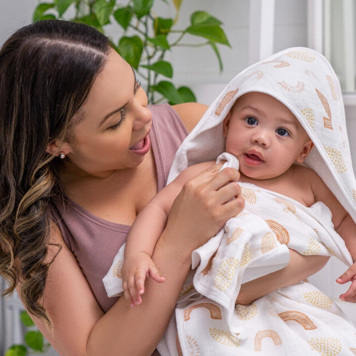 A woman smiles at a baby wrapped in an aden + anais boutique 100% cotton oversized muslin towel adorned with colorful patterns. The baby gazes at the camera, while a green plant stands gracefully in the background.