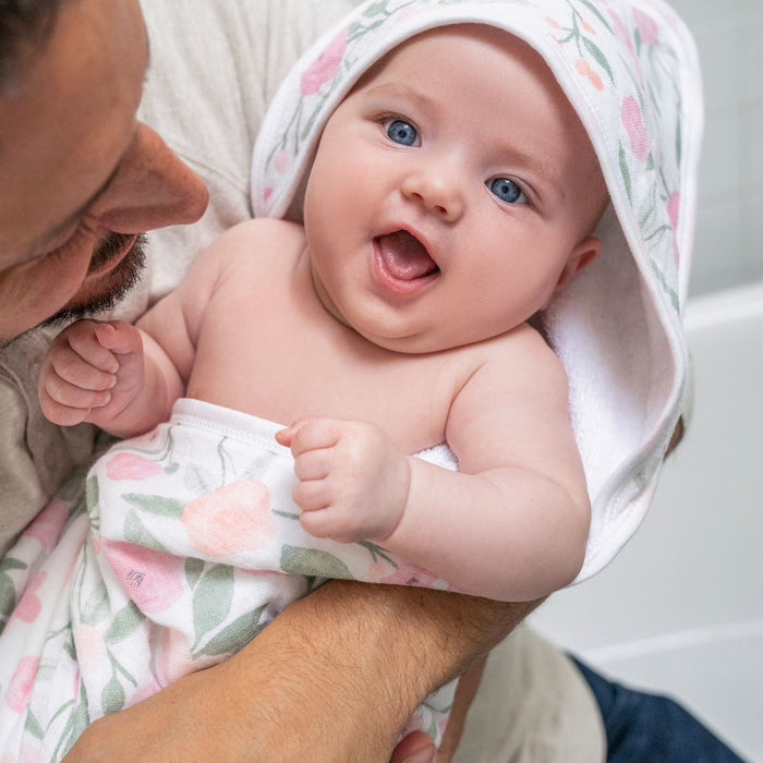 An adult holds a baby wrapped in a boutique 100% cotton oversized muslin-backed towel + washcloth set from aden + anais. The baby, smiling with bright eyes, is cozy in the colorful hooded towel as plush fabric enhances warmth, creating a scene filled with joy and loving gazes.