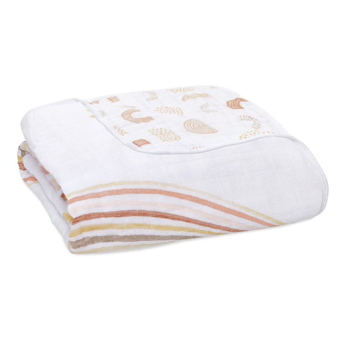 The aden + anais boutique cotton muslin dream blanket is a soft, plush blanket made from breathable cotton muslin. It features pastel rainbow patterns in peach, pink, and beige on a white background, creating gentle, soothing curves.