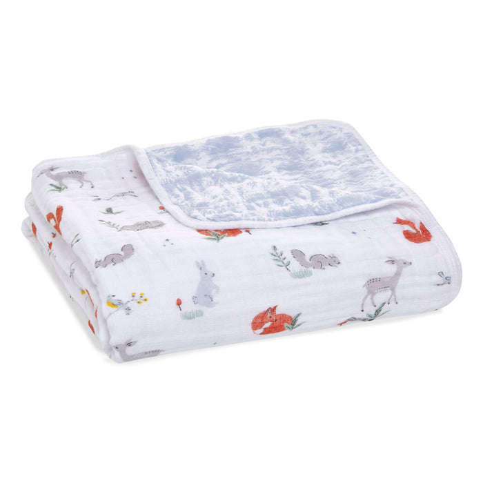 The aden + anais boutique cotton muslin dream blanket displays a charming woodland animal print featuring foxes, squirrels, and rabbits on a white backdrop. Made from breathable cotton muslin, it has a light blue underside with a delicate white pattern, ideal for nurturing cozy dreams.