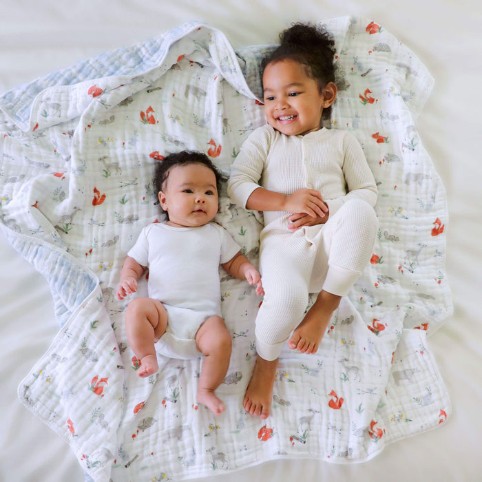 Two young children relax on aden + anaiss soft, colorful boutique cotton muslin dream blanket featuring animal prints. The baby in a white onesie gazes upward, while the other child smiles, stretching their legs in light pajamas made from breathable cotton muslin. The setting features a light surface.