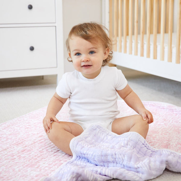 A smiling toddler sits on a cozy cloud of the aden + anais boutique cotton muslin dream blanket in soft pink and light purple in a warm nursery. Clad in a white onesie, theyre surrounded by the gentle charm of a wooden crib and white dresser, wrapped in cotton muslin comfort.