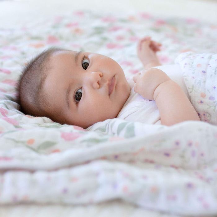 A baby rests on an aden + anais boutique cotton muslin dream blanket with a floral pattern, gazing at the camera. Dressed in white, the infant is enveloped in a breathable and cuddly atmosphere, ensuring a cozy and gentle setting.
