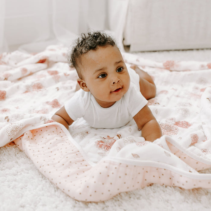 The baby, lying on an aden + anais boutique cotton muslin dream blanket made from 100% cotton, looks curious with parted lips. Surrounded by soft white and pink patterns, the breathable setting feels cozy and bright. Curly hair tops off the serene scene as the little one explores their world.