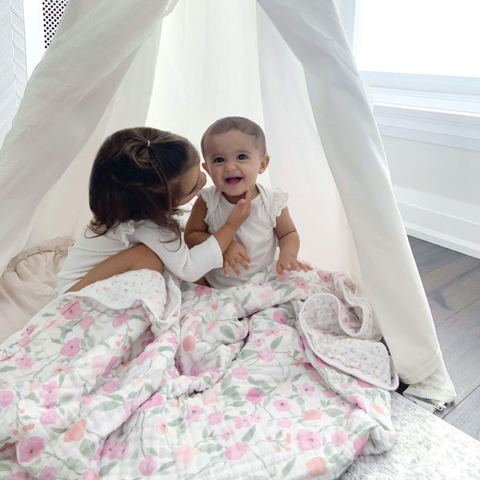 A young girl and a baby sit under a white canopy on a wooden floor, wrapped in an aden + anais boutique cotton muslin dream blanket. The extra soft, 100% cotton muslin provides a breathable and cuddly embrace as the girl gently touches the babys face, both smiling in the cozy setting.