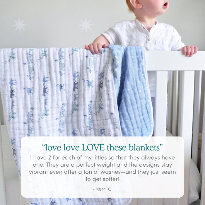 A baby stands in a white crib holding the rail, with two aden + anais boutique cotton muslin dream blankets—one white with blue dinosaurs and one light blue—draped over the side, highlighting their cozy comfort. A customer review is shown below.