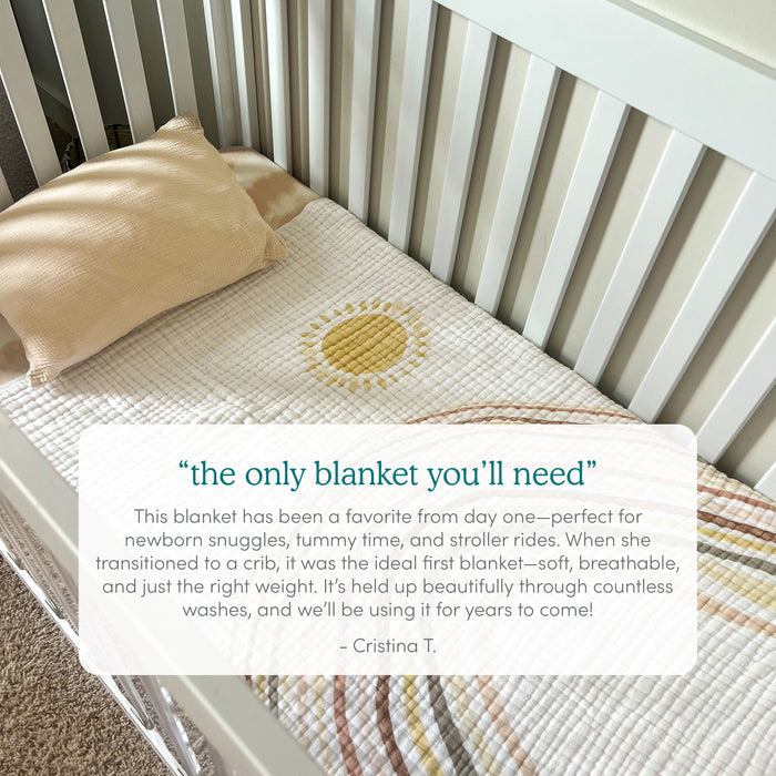The aden + anais boutique cotton muslin dream blanket is nestled in a white crib with a simple pillow, displaying a sun and rainbow design. Perfect for newborns and beyond, it offers softness, breathability, and durability as praised by Cristina T.