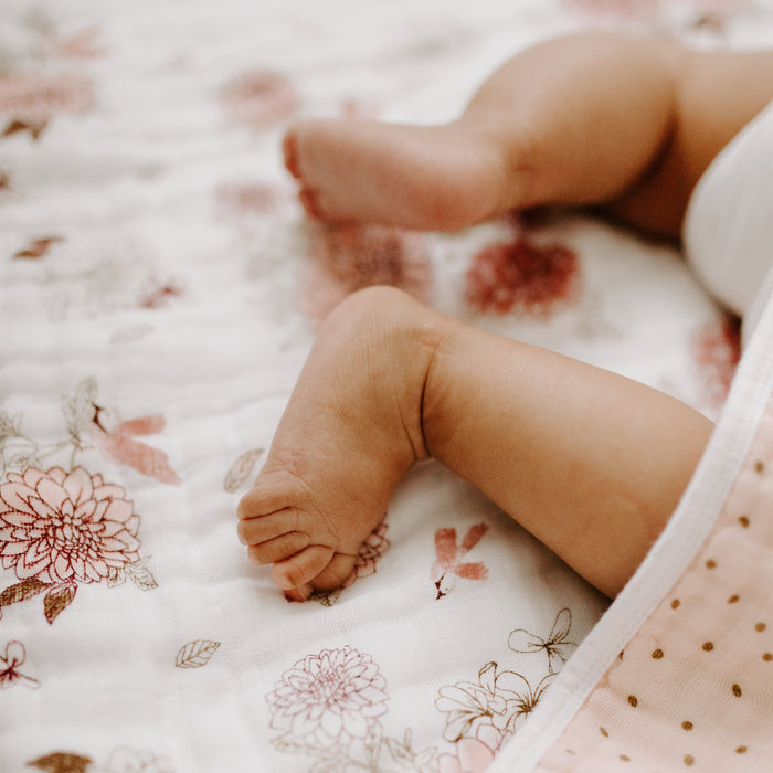 A close-up shows a babys legs on an aden + anais boutique cotton muslin dream blanket. One leg is bent, with relaxed feet partially covered by the pink and white blanket adorned with tiny polka dots and soft floral designs.