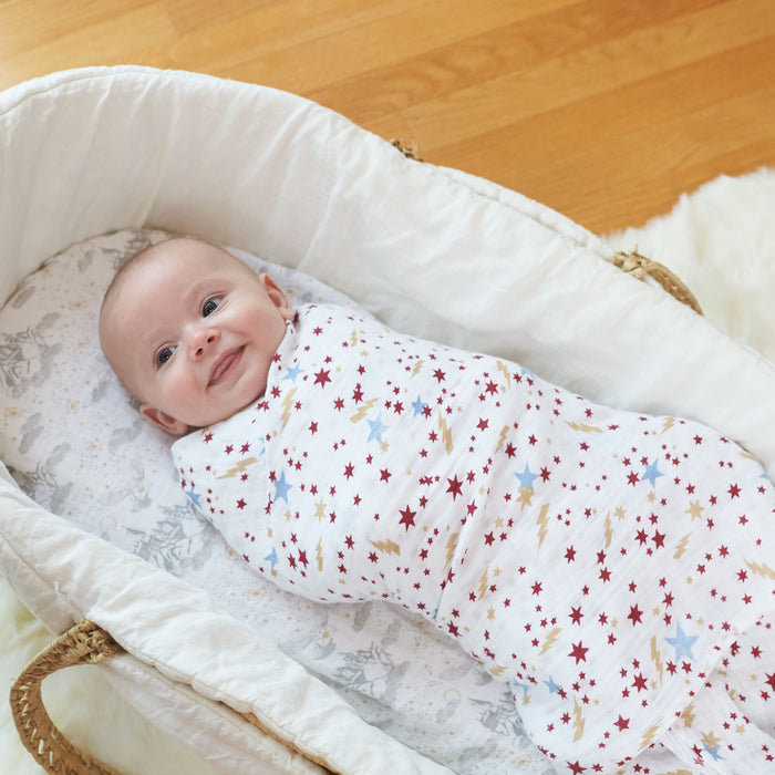 A smiling baby is snugly wrapped in an aden + anais boutique iconic 100% cotton muslin swaddle blanket with red stars, nestled in a cushioned basket on a wooden floor. The baskets soft lining features delicate animal prints, enhancing the charm of this magical scene.