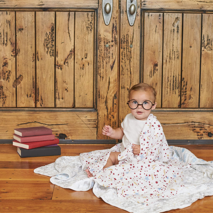 A baby, resembling Harry Potter with round glasses, sits on a wooden floor by a rustic door wrapped in an aden + anais boutique iconic 100% cotton muslin swaddle adorned with red and blue stars, with a stack of books nearby completing the charming scene.