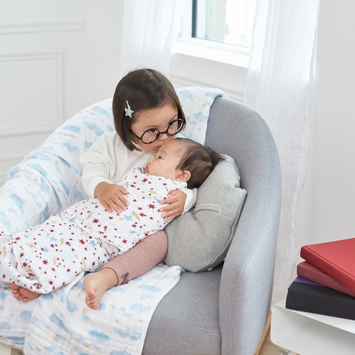 A young child with glasses, resembling a Harry Potter character, sits on a gray armchair holding a baby wrapped in the boutique iconic cotton muslin swaddle by aden + anais. They are in a cozy room with a window and books stacked nearby.