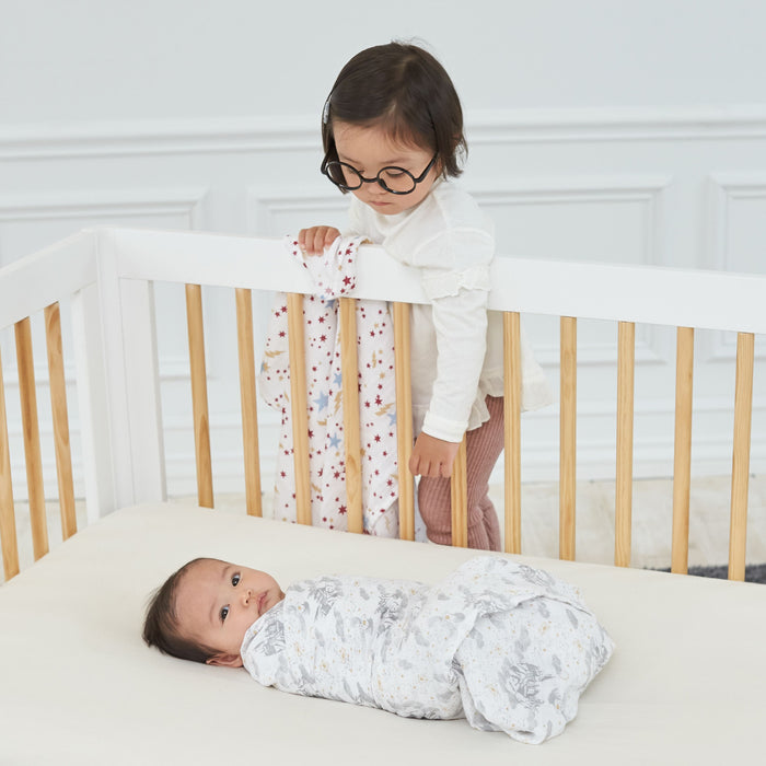 In a white, minimalist room, a toddler with glasses reminiscent of young Harry Potter stands inside a crib, looking down at a baby wrapped in the aden + anais boutique iconic 100% cotton muslin swaddle. The toddler holds a floral blanket draped over the crib rail.