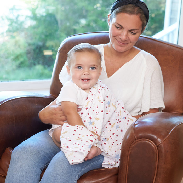 A woman sits on a brown leather chair, holding a smiling baby wrapped in an aden + anais boutique iconic 100% cotton muslin swaddle blanket adorned with colorful stars, with a window view of lush greenery in the background.