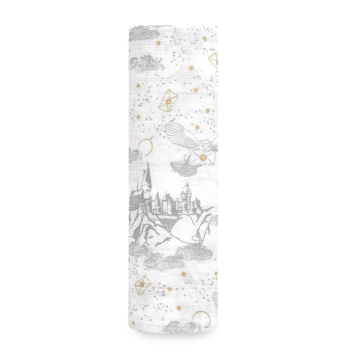 The aden + anais boutique iconic 100% cotton muslin swaddle blanket features rolled-up white fabric with subtle gray designs of castles, magical symbols, and flying creatures. Gold stars and crescent moons add whimsy, reminiscent of a Harry Potter™ adventure.
