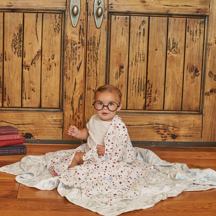 A baby in round glasses sits on a wooden floor wrapped in an aden + anais boutique iconic cotton muslin dream blanket, in front of rustic doors with a stack of books nearby, creating a cozy, Harry Potter-esque whimsical scene.