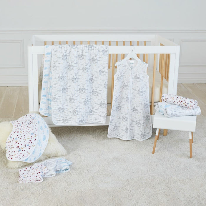 The nursery includes a white crib with wooden slats, featuring aden + anais boutique iconic cotton muslin dream blanket in floral and polka dot patterns. A zip-up baby garment hangs on the crib, while a stool holds folded blankets. A white rug and plush item sit on the beige floor.
