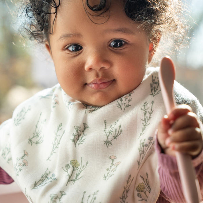 A baby with curly hair wears an aden + anais boutique silky soft floral burpy bib, holding a pink spoon. The 2-in-1 bib ensures extra absorbency as the baby gazes gently at the camera. Soft daylight filters through a window in the background.